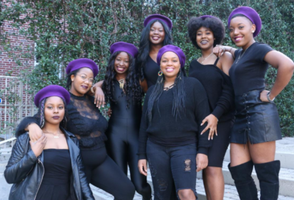 7 Black women in all black clothes and purple berets pose in front of greenery
