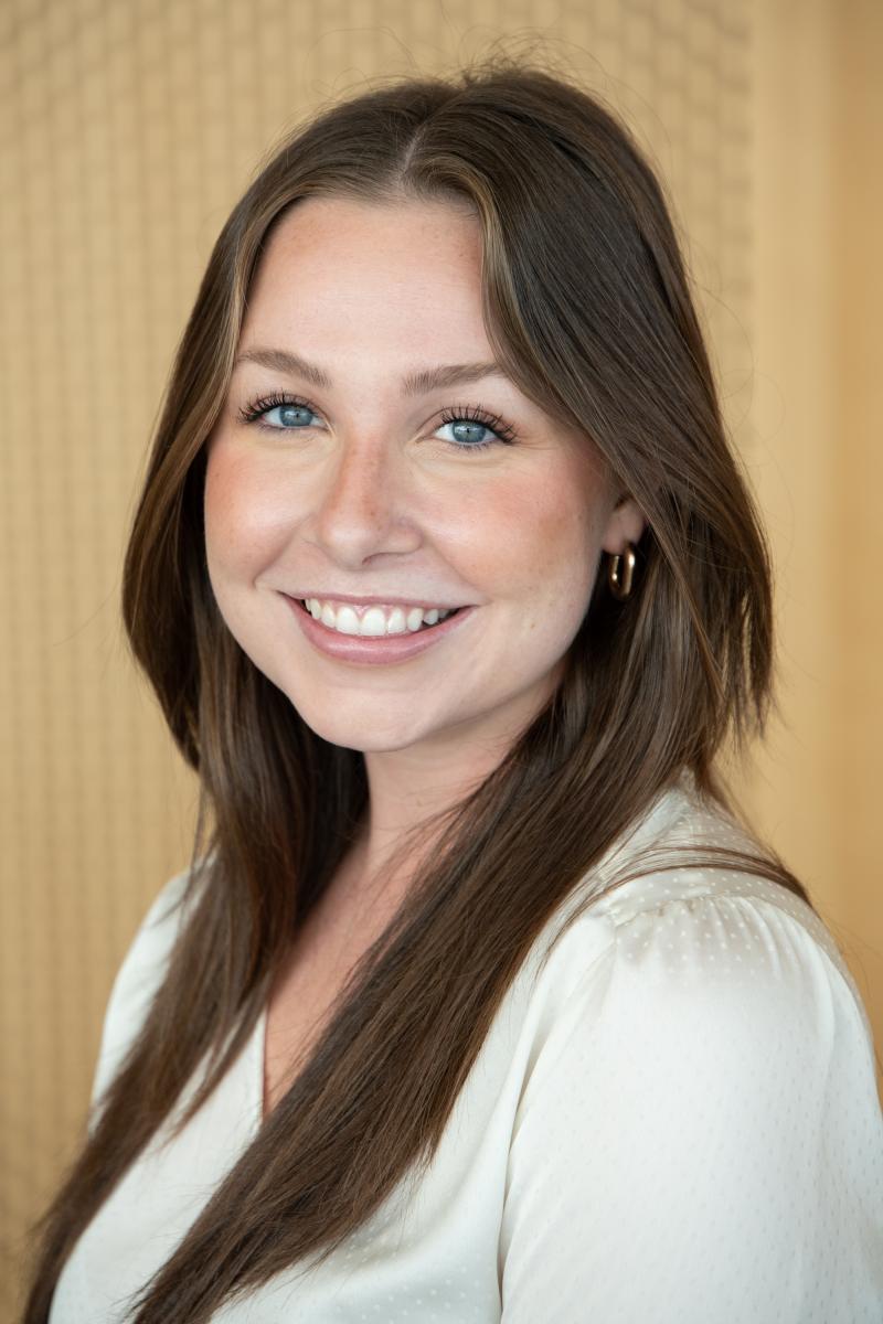 Headshot of Katie Elder, she stands in front of a wood panel wall and is wearing a white shirt 