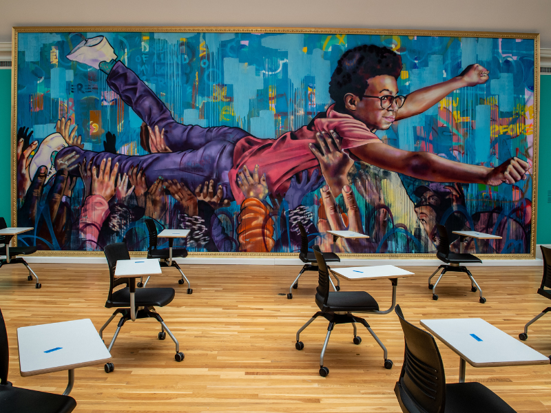photo of classroom in art gallery, features painting of a young person being carried by a group people in a Superman pose