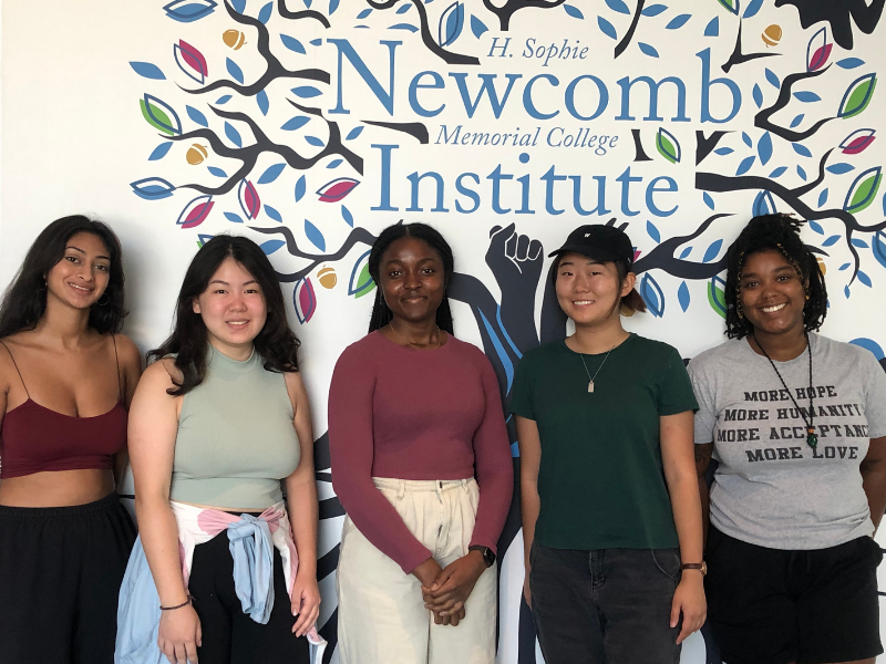 Group of five students stands in front of a mural with an oak tree with the words "Newcomb Institute"