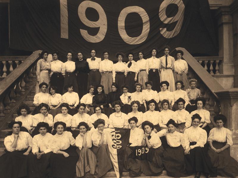 Photo of the 1909 students of Newcomb College posing on the stairs with a larger banner behind them reading "1909"