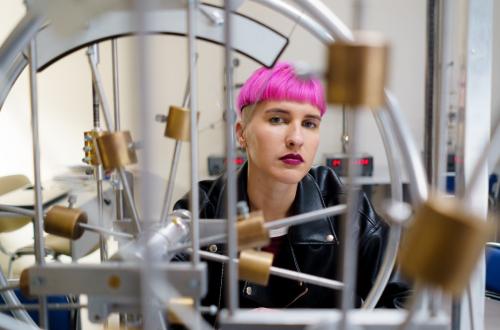 Courtesy of https://apscuwipattulane.com/ A person with pink hair stands behind some metal stuff