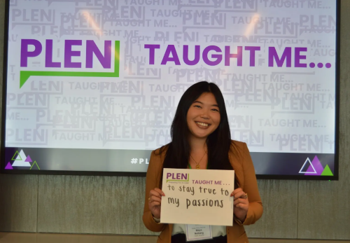 An Asian girl stands in front of a projector screen that says "PLEN taught me" holding a sign that says "PLEN taught me to stay true to my passions"