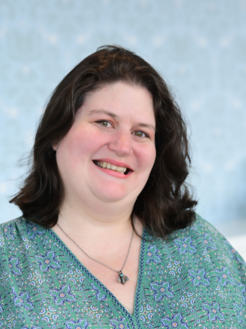 White woman with dark hair and green shirt and bee necklace smiles for the camera.