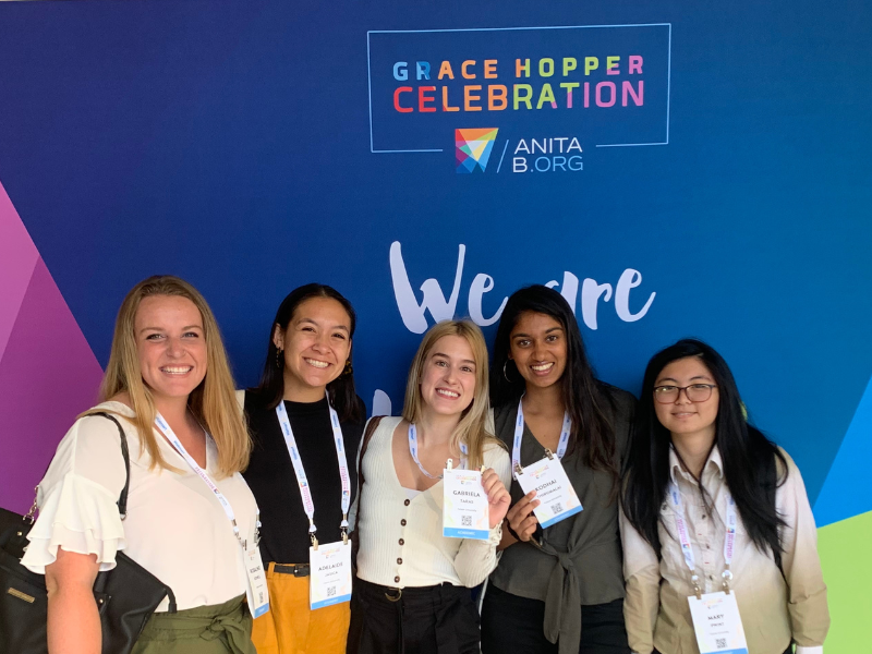 Students stand in front of a colorful backdrop at he Grace Hopper Conference